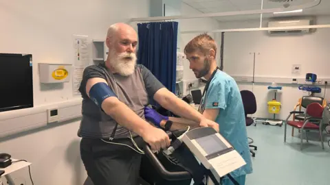 Graeme Harrison at University Hospital Southampton's Clinical Research Facility he is on an exercise bike with a member of medical staff watching him. Graeme has a long white beard and wears a grey t-shirt as well as as heart monitoring equipment.