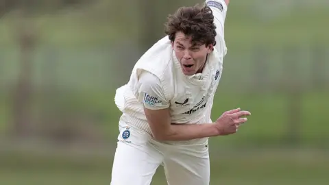 Henry Brookes crouched forward with his right arm held up in the air behind him after he bowls a ball during a friendly for Middlesex