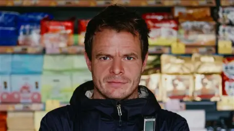 Dan Kelly Dan Kelly looking at the camera with a slight smile. A full supermarket shelf is visible behind him.