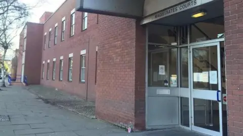 A red-bricked double-storey building with a large glass door. Above the door is a sign that reads: "Magistrates' Courts". There are paving slabs leading up to the entrance. 
