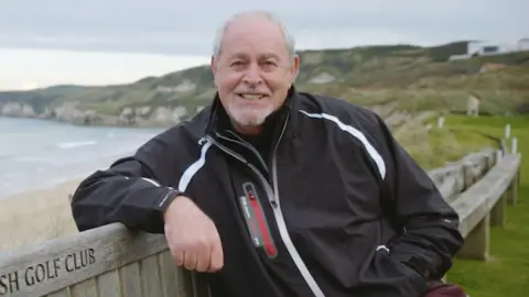 A man with short white hair smiles at the camera. He is wearing a black jacket and sitting on a bench outside. Behind him is a beach.