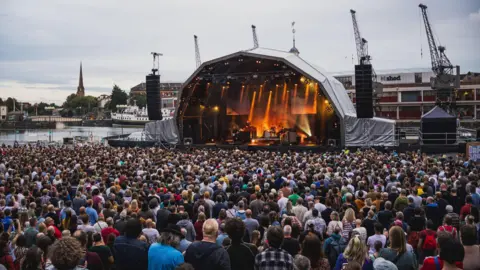 A large crowd is seen in front of the stage at Bristol Sounds 2025 during the Supergrass concert. The picture is taken from the back of the crowd and behind the stage the harbourside cranes are visible