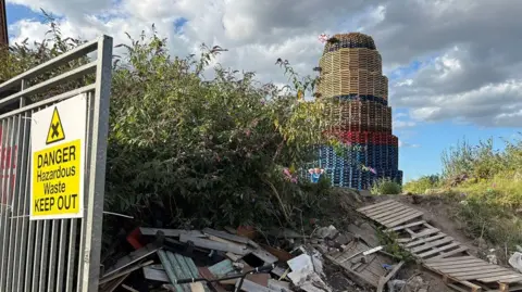 Bonfire at south Belfast site, it has red and blue and brown pallets and is surrounded by debris and old pallets 