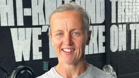 Laura Payne is smiling at the camera, standing in a gym with a rowing machine behind her. She has a grey shirt and swept back grey hair.