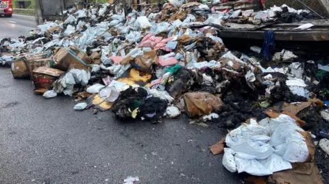 T J Cottis Transport Ltd Lots of burnt and damaged packages on a road. There are lots of bags, mostly paper and plastic. They are piled up beside a lorry. 