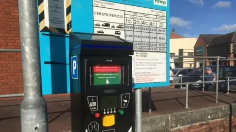 LDRS A black parking meter, with a sign listing charges behind it and a lampost to the left.