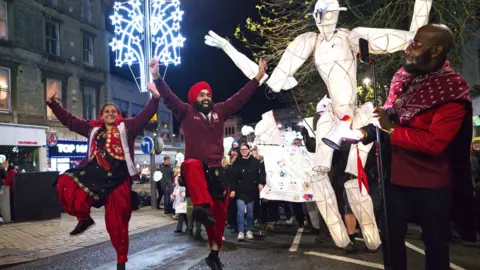 A man and woman are dancing with their arms in the air, while a man looks like he is recording the peformance on a phone which is on a stand. Behind them is a large sculptured figure, which is lit up, and a street light decoration which is also lit up. The performance is in the street in the evening and there are people watching and some shops in the background. 