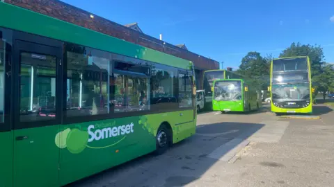 BBC A green, single-decker bus is parked up at a depot. It reads 'the buses of Somerset on the side. In the background are two double-decker buses and another single-decker bus. They are all green. 