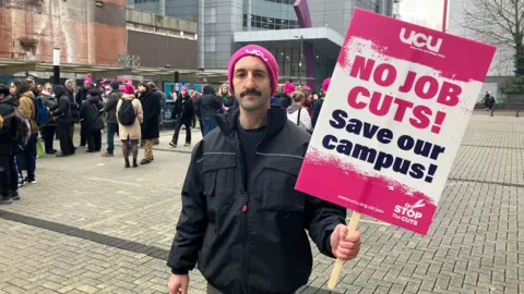 Alex Stevanovic/BBC A man wearing a black jacket and a pink UCU-branded hat. He is holding a placard saying "No Job Cuts"" and "Save our campus!" There are other demonstrators standing in the background.