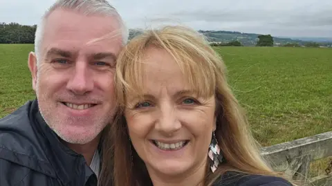 Cancer Research UK handout Justin (left) with grey hair and stubble wears a blue jacket hugs Gail Hardy (right) who has long blond hair and dangly bronze and silver leaf style earrings. They are standing in front of a field and are both smiling.
