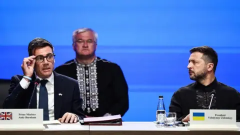 PA Media Andy Burnham adjusts his spectacles on the left of the image as Ukrainian president Volodymyr Zelensky looks across at him. The pair are on a stage and have name plates and folders in front of them.