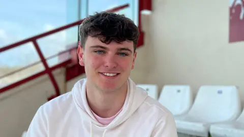 Ollie smiles at the camera as he sites on the football stands and he's wearing a white hoodie with a pink T-shirt underneath.  He has short black hair and blue eyes.