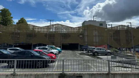 Ben Parker/BBC An open-air car park is in the foreground with multiple cars parked in spaces. In the background is a multi-storey car park with a yellow grille-type facade.
