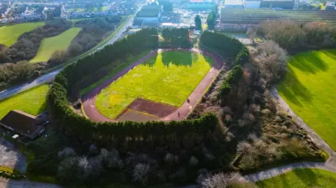 John Pearce An aerial photo taken from a different angle of the Whitchurch race track. There is a green field in the middle of the running track and the track is surrounded by thick green trees and bushes. Some houses can be seen in the distance.