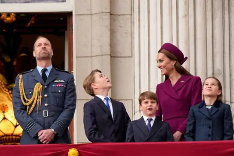 PA Media William, Prince of Wales looks up to the sky while Catherine, Princess of Wales laughs and their three children look up to the sky as they stand on the balcony of Buckingham Palace.