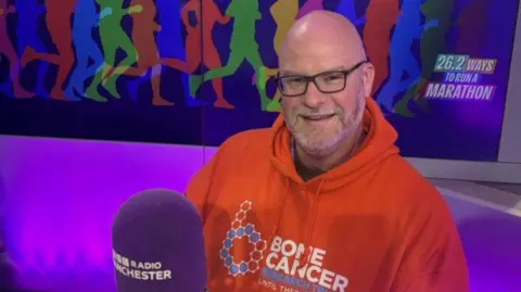 Peter Lloyd, who is wearing a bright orange Bone Cancer Research Trust hooded top, is sitting in the BBC Radio Manchester studio. There is a BBC-branded purple microphone on a desk in front of him. 