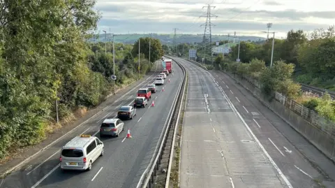 A line of traffic on the Sydenham Bypass as long as the eye can see. The other lane is closed and the road on the other side is closed too. It is a sunny day. 