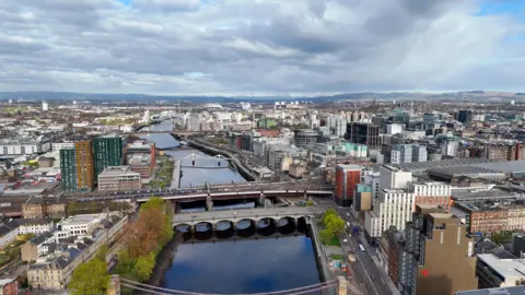Aerial view of Glasgow city centre showing the River Clyde, multiple bridges, railway lines and modern office buildings beneath dramatic clouds stretching across the skyline.