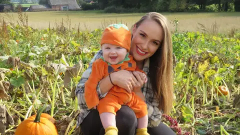 Emma Powell Emma and Leo in a pumpkin patch . Leo has an orange jumper and hat and is sitting on mum's knee with a big smile