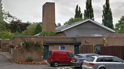 York Crematorium near Bishopthorpe Road, with roses growing on the building and vehicles parked to one side. The crematorium's chimney is prominent. 