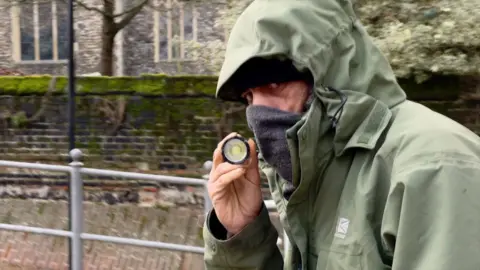 Alex Dunlop/BBC Christopher McEwen looks directly at the camera, wearing a green jacket with the hood up. He has a scarf covering his nose and lower part of his face. He is holding up and pointing an unlit torch at the camera. 