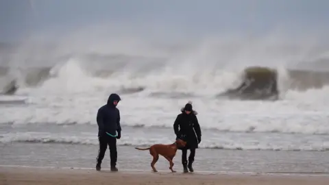 Dog walkers in the wind on Tynemouth Longsands on the North East coast of England. Large waves have formed in the background.