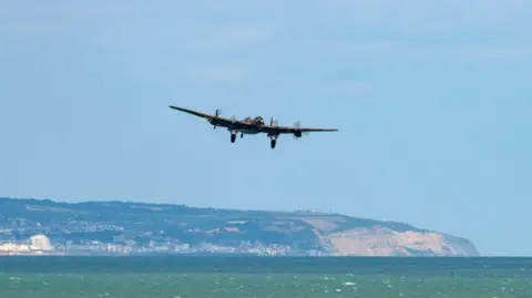 Claire Hartley An old WW2-era bomber plane flys low over the ocean. 