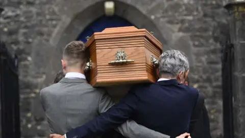 The brown coffin of Lord Mount Charles, being carried at St Patrick's Church of Ireland in Slane. On the left is a man with fair hair and a grey suit, on the right, a man with grey hair and a dark blue suit.