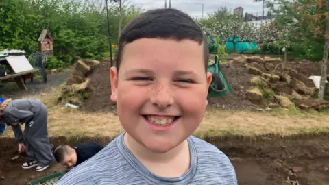 A child with short brown hair smiles at the camera. He wears a grey t-shirt and stands in front of a large pile up dug-up brown mud. Two children can be seen over his right shoulder. 