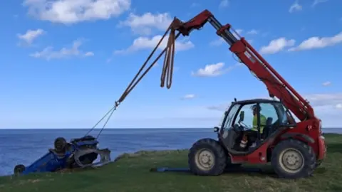An upturned blue Peugeot being hauled to safety from a cliff by a red tractor. A recovery lorry is parked behind the tractor. White clouds are dotted in the blue sky. The blue sea can also be seen from over the cliff. 