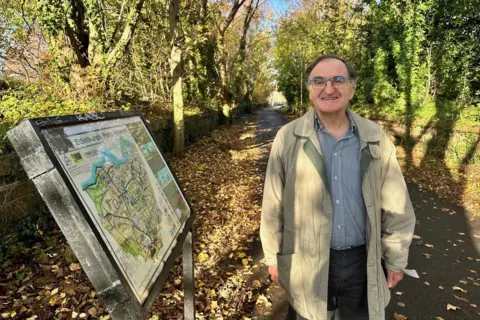Lawrence Marshall is standing on the Roseburn Path beside a board showing all the defunct rail routes in Edinburgh. The path is lined with trees and there are yellow and brown leaves on the ground. He is wearing glasses and a long, green jacket and has brown receding hair. He is smiling at the camera.