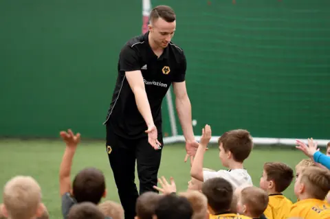 The Wolves Foundation Diogo Jota wearing a black Wolves kit stands on his pitch in front of crowds of children sitting down - many wearing Wolves kits. His hands extend down towards theirs.