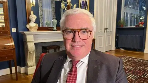 Carl Decaluwé looks at the camera and smiles. He is wearing a dark suit, white shirt and red and white spotted tie. Behind him is a large fireplace and dark blue painted walls.