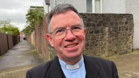 A man smiles at the camera. He has large, thin rimmed glasses and wears a priest's clothes - blazer, shirt, clerical collar. 