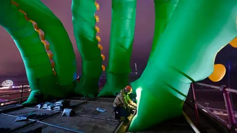 VisitBlackpool Six huge inflated tentacles being installed on a rooftop at night by a workman who is dwarfed by them. The tentacles are green with yellow nodules and they rise up into the air