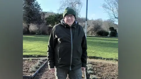 GL Communities Roy Kellett stands in the Matson Rose Garden. Roy is wearing a dark green beanie hat, black framed glasses, a black zip-up waterpoof jacket and khaki coloured trousers. There is a lawn behind him with a row of tall trees and a bright blue sky with a couple of whispy light clouds. Just behind Roy's legs are mostly empty flowerbeds with soil visible and a few shrubs.