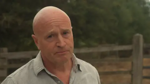 A head and shoulders image of a man wearing a grey shirt. He is standing in front of a wooden fence and some trees. He is looking into the camera. 