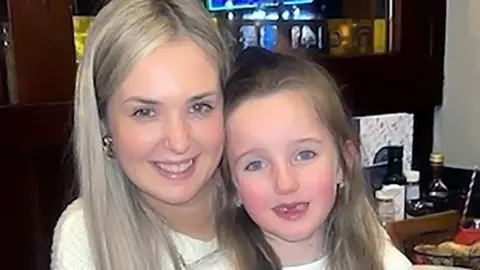 Rebecca Hill in a white jumper with long blonde hair and gold earings, cuddles daughter Arianna, with long hair and two missing front teeth, in a close up photo of their faces