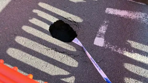 A sinkhole is in the middle of the photo, with blue-and-white police tape going from the bottom right of it. 
