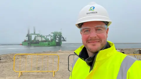 Peter is wearing a white hard hat with the logo and name of his employer on it. He is wearing a high visibility jacket. Behind him is a large green ship.
