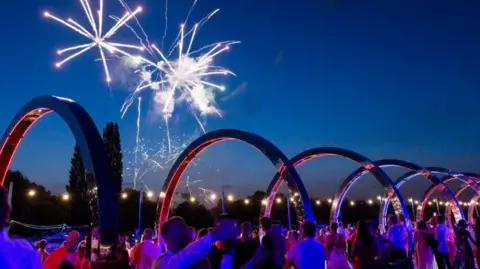 James Robinson. The Henley Festival at night with crowns under a arch on the river bank watching fireworks.