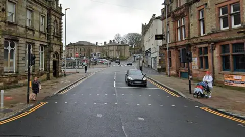 View of Church Street in Darwen town centre. There is a car waiting at traffic lights and a woman wearing a white top is waiting to cross the road with a child who is in a buggy.