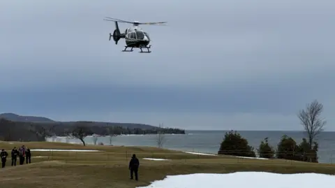 A helicopter seen taking off. There is snow and people standing nearby on grass