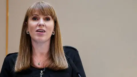 PA Media Deputy Prime Minister Angela Rayner stands at a lectern to address an audience. She has shoulder-length brown hair and wears a black dress.