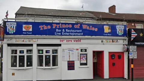 The outside of pub with a white building and a red door. It says 'The Prince of Wales' in a blue sign.
