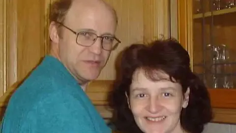 Greg Worrall A man wearing a blue jumper and sporting spectacles stands with a woman who has brown hair, both are looking at the camera