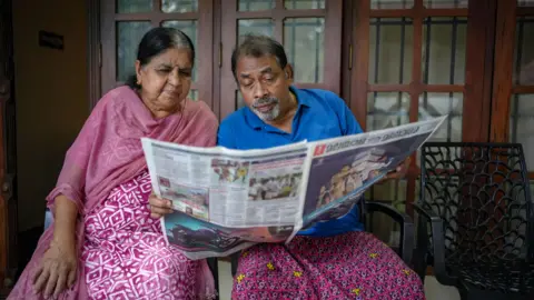 Arun Chandra Bose TO Dominic and his wife sit side by side indoors, holding and reading a newspaper together.