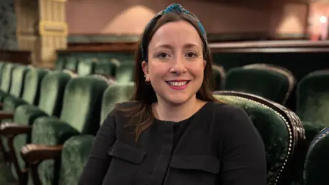 Sarah Wilson-White sits on the green velvet seats of the stalls of the theatre. She has shoulder length brown hair, which is tied back.