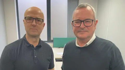 Ashley Riley and Lee Barron stand side-by-side. Riley is in a navy polo shirt with short sleeves and Barron is wearing a navy jumper over a white buttoned shirt. Both men have eye glasses with black frames and are standing in an office environment.