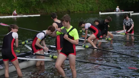 Eight rowers standing shin deep in the River Wear with their vessel. One rower is holding a black triangular fin that has broken off.